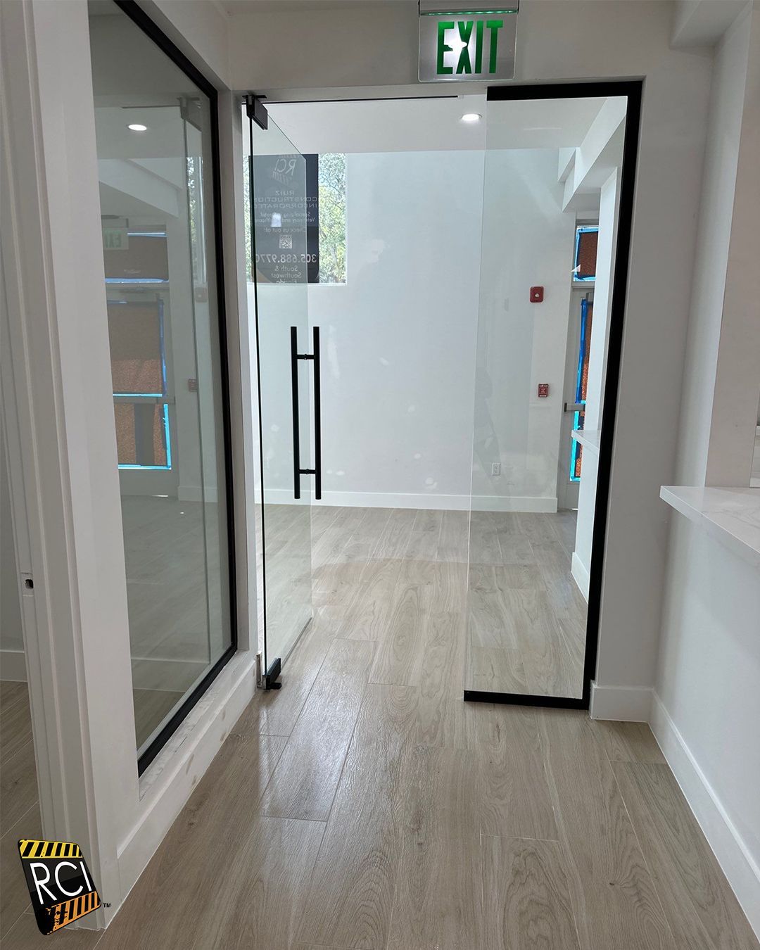 Glass entry doors with black frames and handle, leading into a bright room with wooden floors and exit sign.
