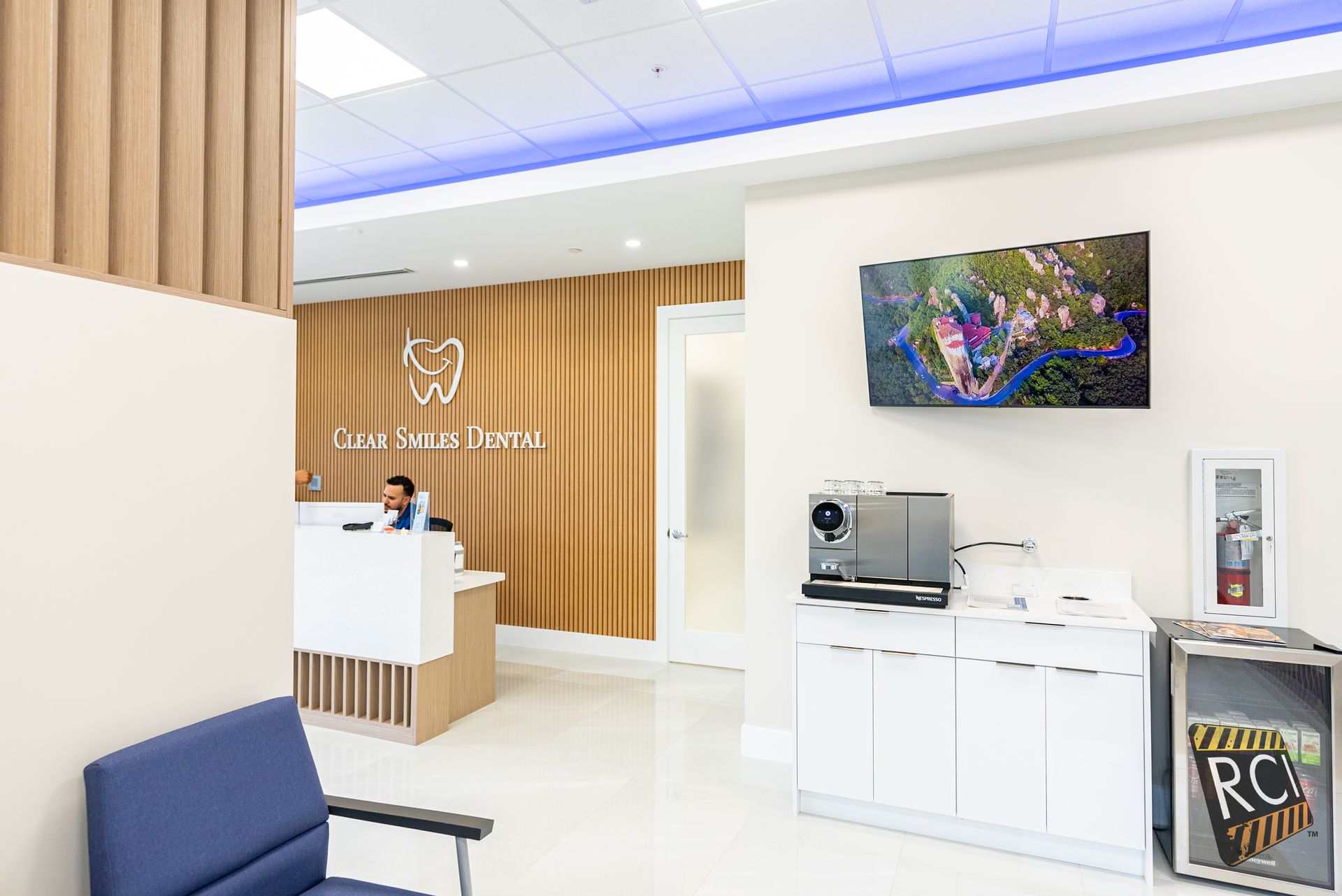 Reception area of a dental office. White and wood paneling, a TV, coffee machine, and a patient sitting in a blue chair.
