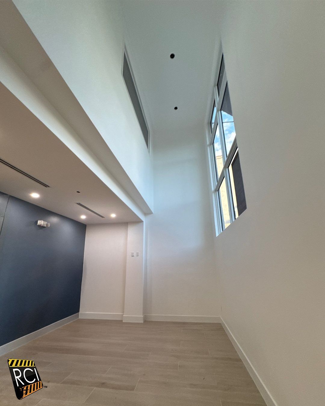 Interior view of a white-walled room with a tall ceiling, natural light from a window, and light wood floors.