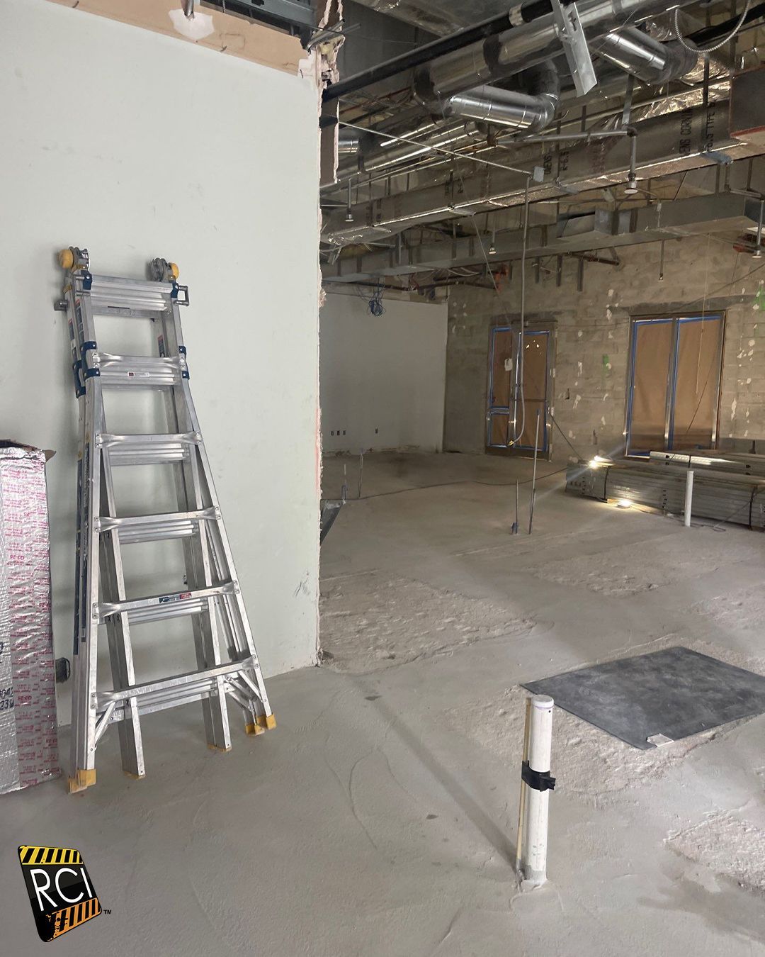 Construction site interior with a ladder against a wall, exposed ceiling pipes, and doorways.