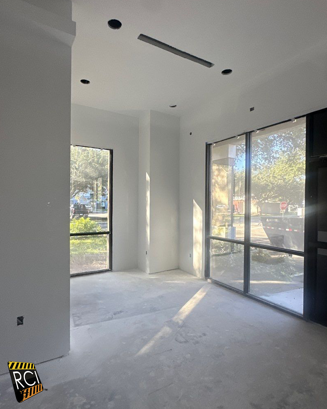 Empty interior room with large windows, unfinished concrete floor, white walls, and recessed lighting.