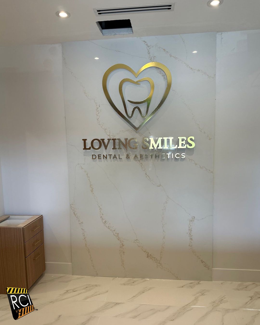 Sign for Loving Smiles Dental & Aesthetics on marble wall. Gold heart-shaped logo with tooth inside, gold text.