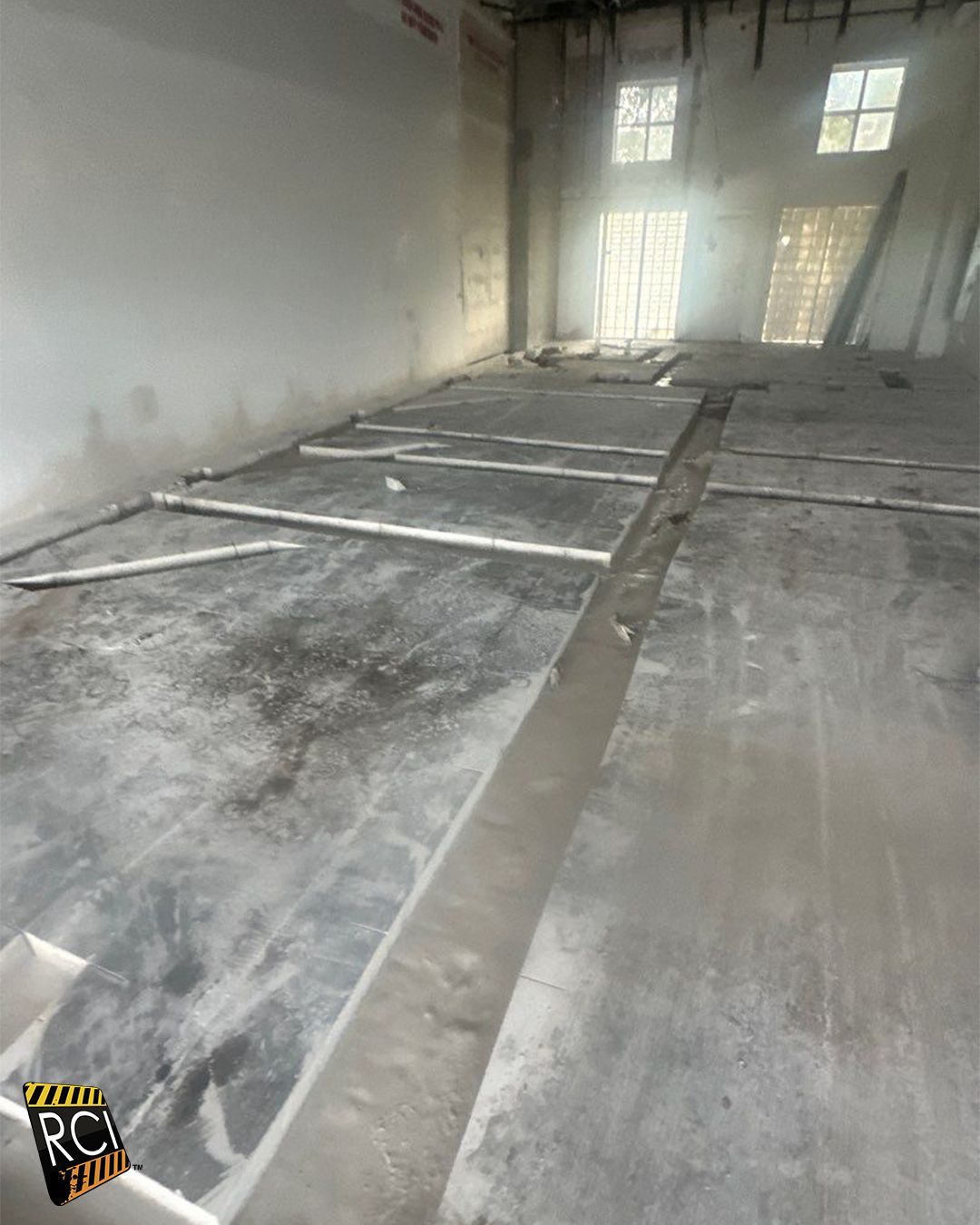 Interior of a room under construction with cut concrete floor and white supports. Windows and light at the end of the room.