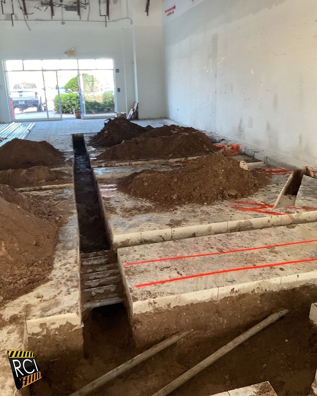 Construction site interior. Trenches dug in concrete floor, piles of dirt, open doorway.
