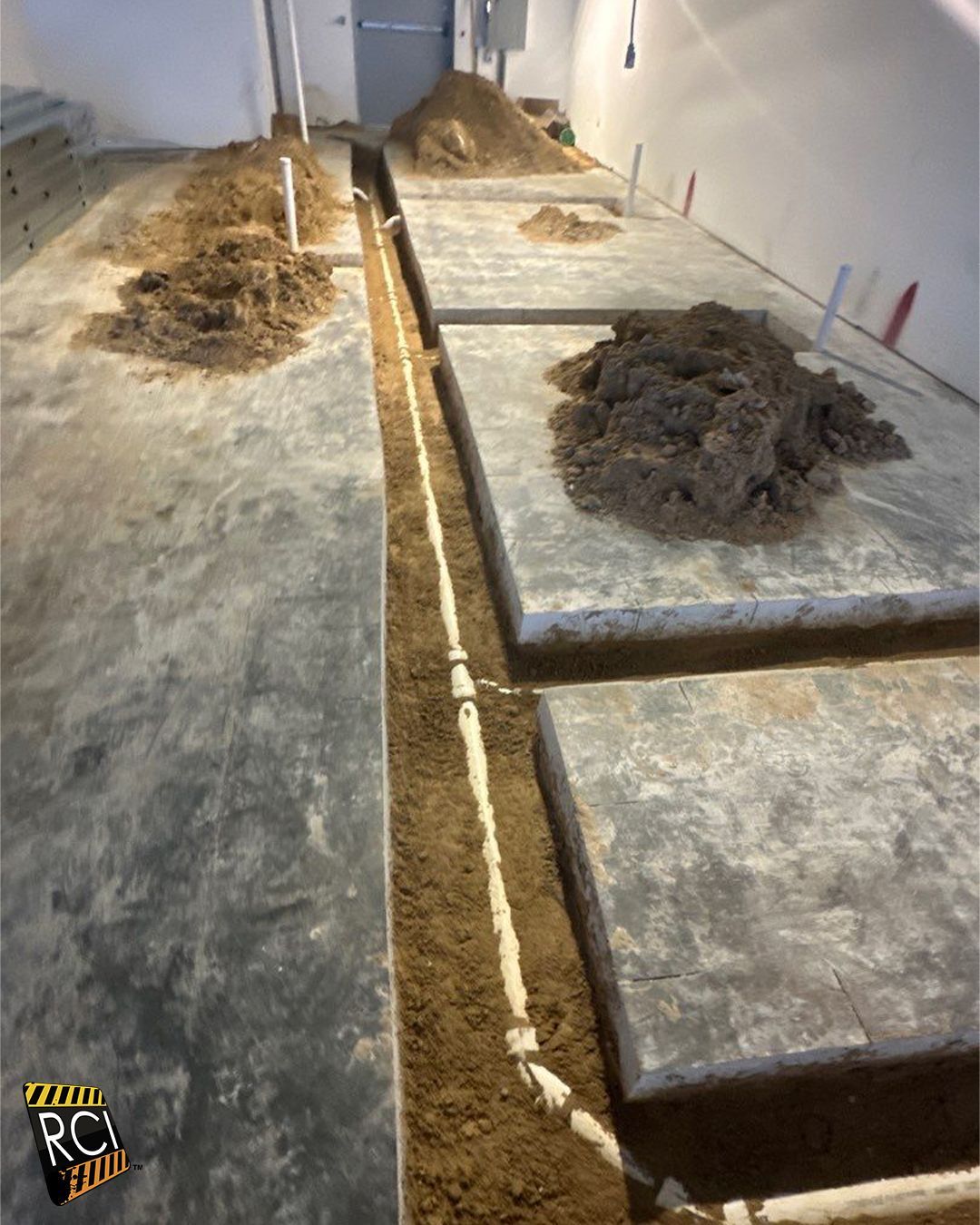Construction site with trenches dug in concrete floor, exposing piping and piles of dirt.