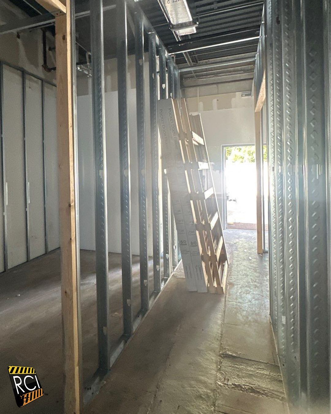 Interior construction: Metal studs form walls; a ladder leans against a partially built wall in a hallway with natural light at the end.
