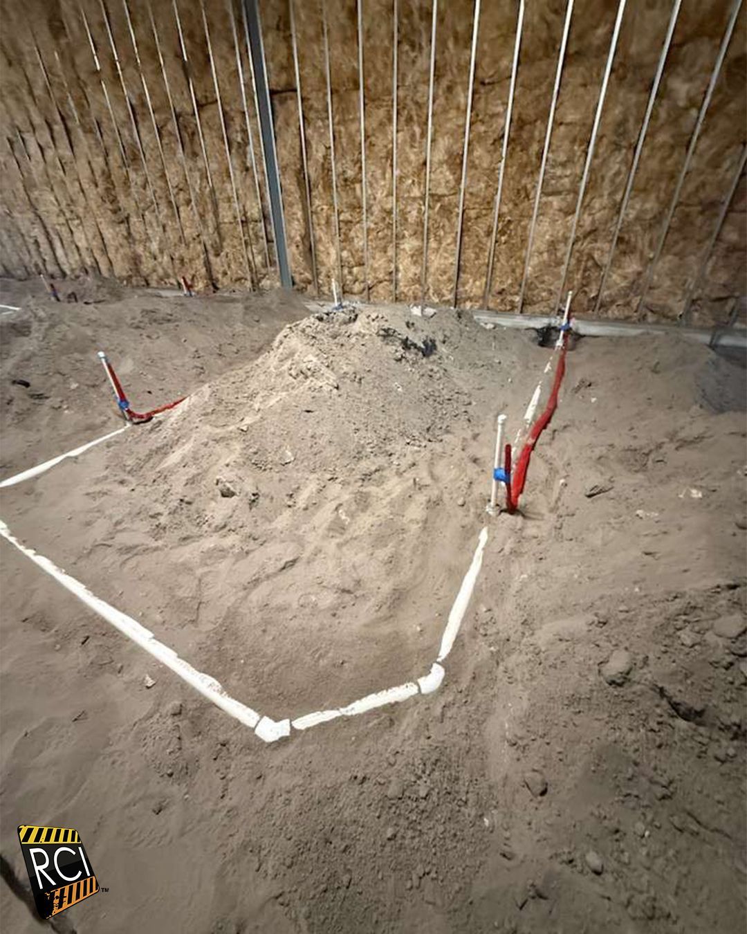 Construction site: white pipes outlining a corner on a dirt floor. Red and blue pipes emerge. Wood wall background.