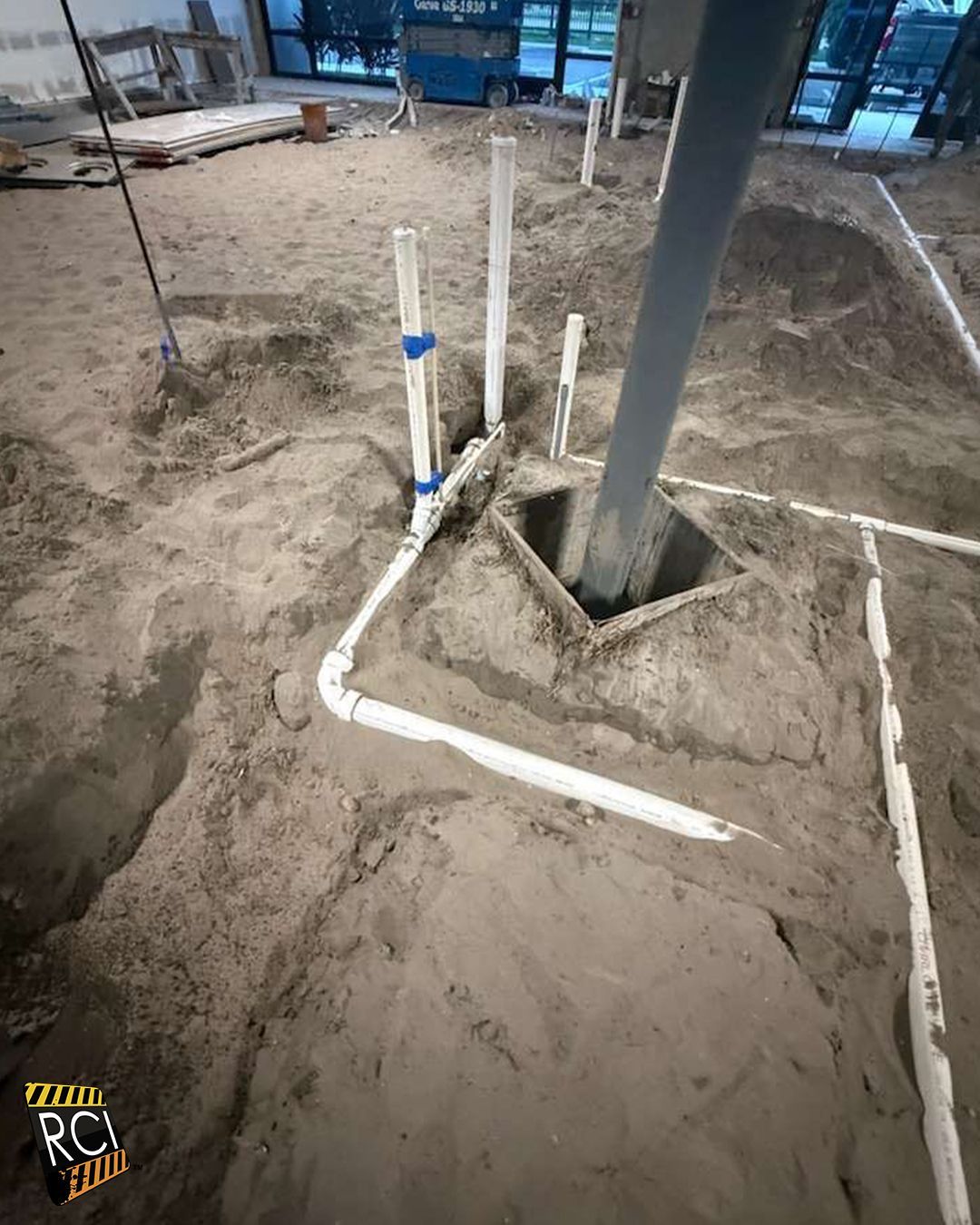Plumbing pipes in a dirt floor; a metal pole descends into a square hole. Construction site.