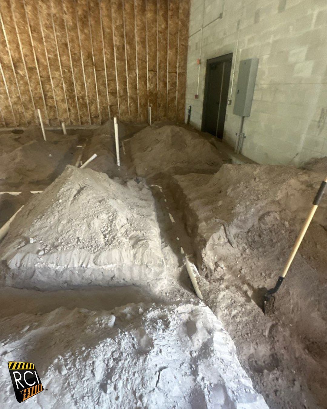 Interior construction site: trenches dug in dirt floor, wood-framed wall, plumbing pipes, concrete block wall, shovel.