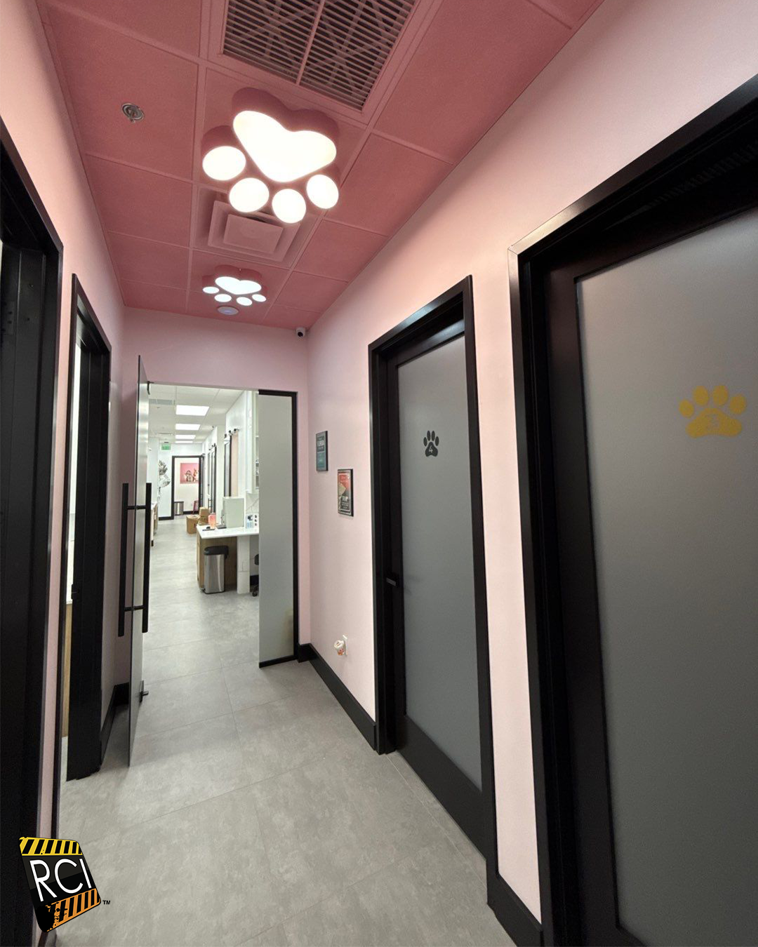 Hallway with pink ceiling, paw-shaped lights, and black-framed doors. Gray flooring, leading to a bright room.