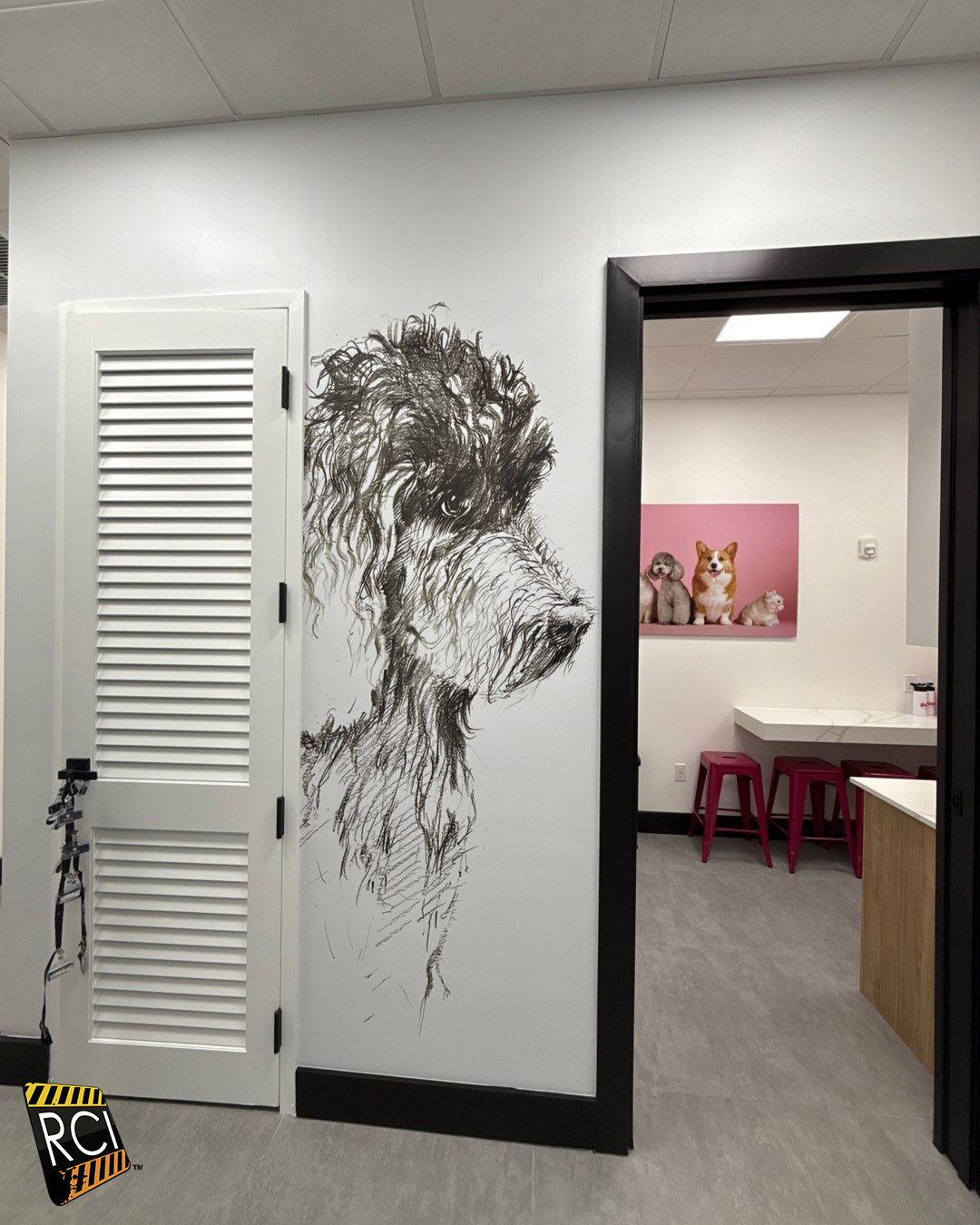 A poodle art piece on a white wall next to a white door. A room with dogs is visible through an open door.