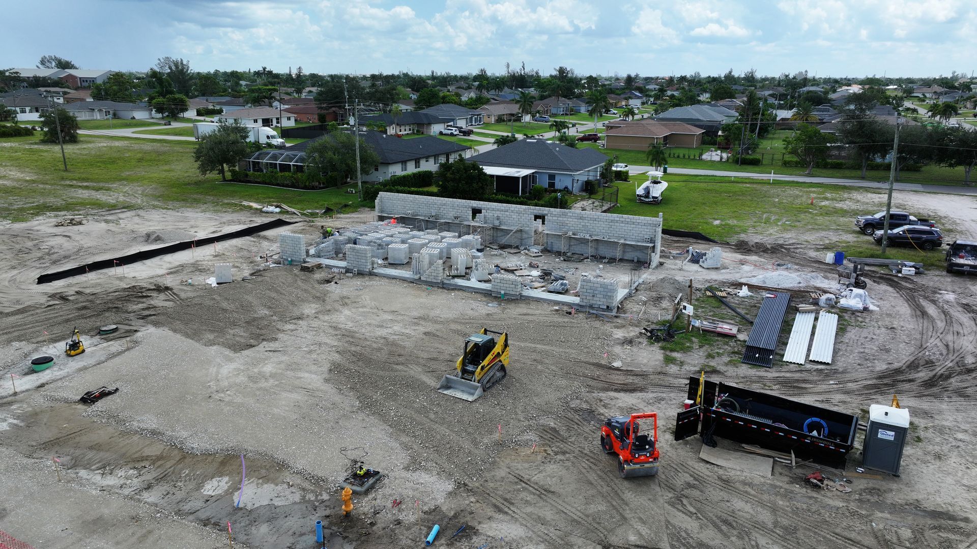 Construction site with machinery, concrete blocks, and houses in the background on a cloudy day.