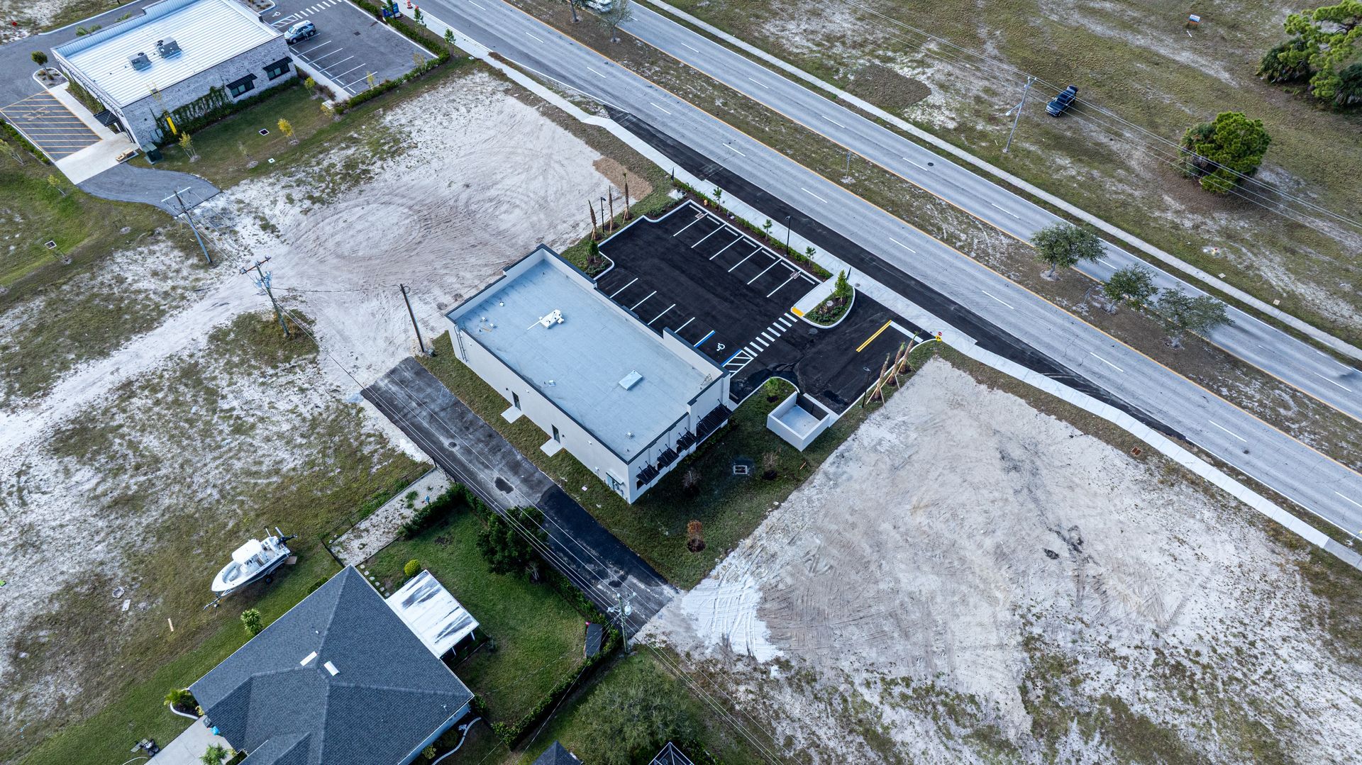 Aerial view of a modern building with a dark roof and a paved driveway, surrounded by empty lots and a road.