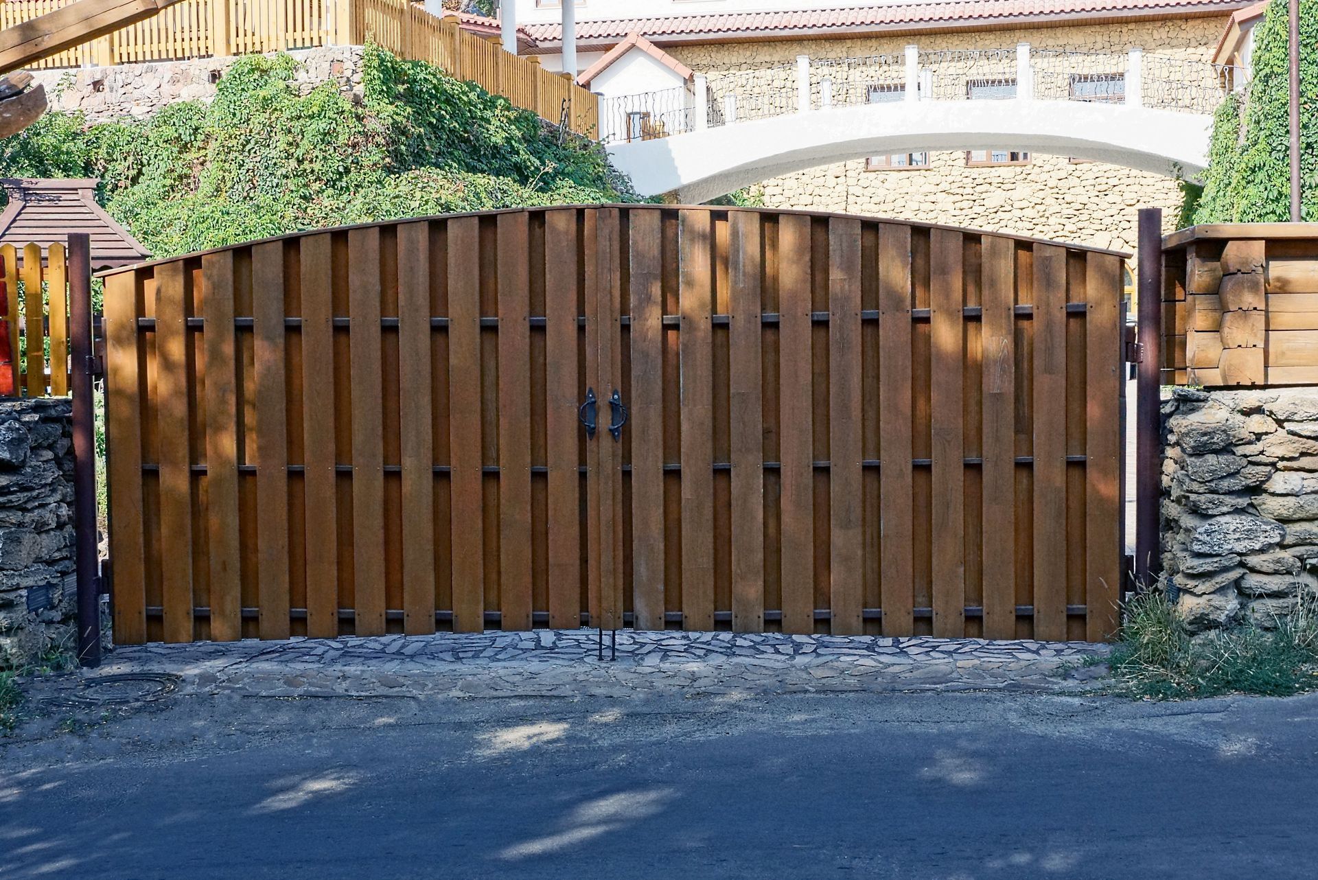 A wooden double gate with a curved top is set between two stone pillars in front of a house with a stone archway.