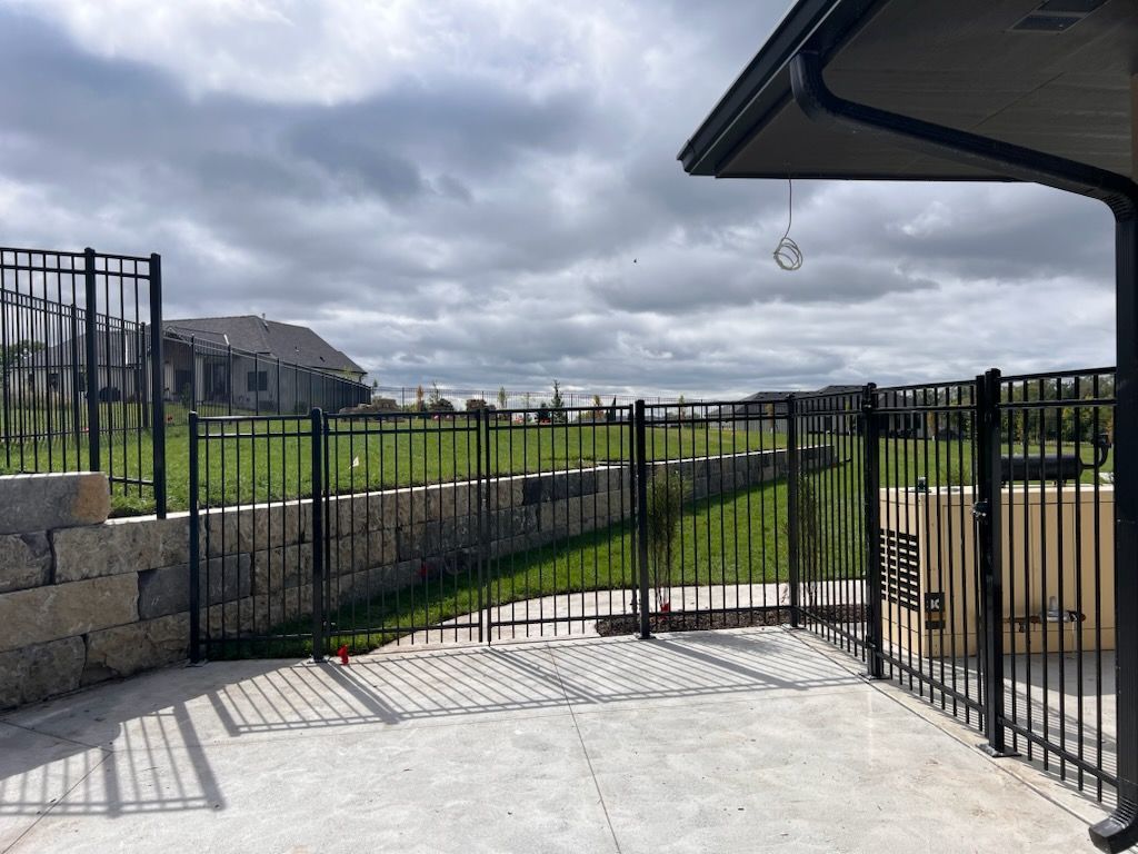 Concrete patio with black metal fence and gate opening to a grassy yard under a cloudy sky