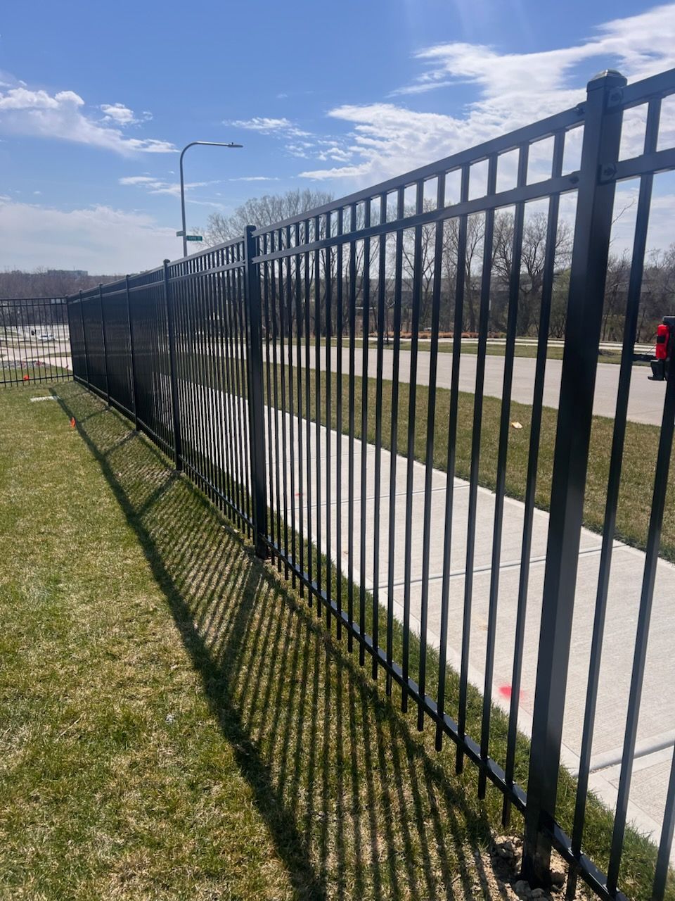 Black metal fence along a sidewalk and road on a sunny day, with grass and long shadows.