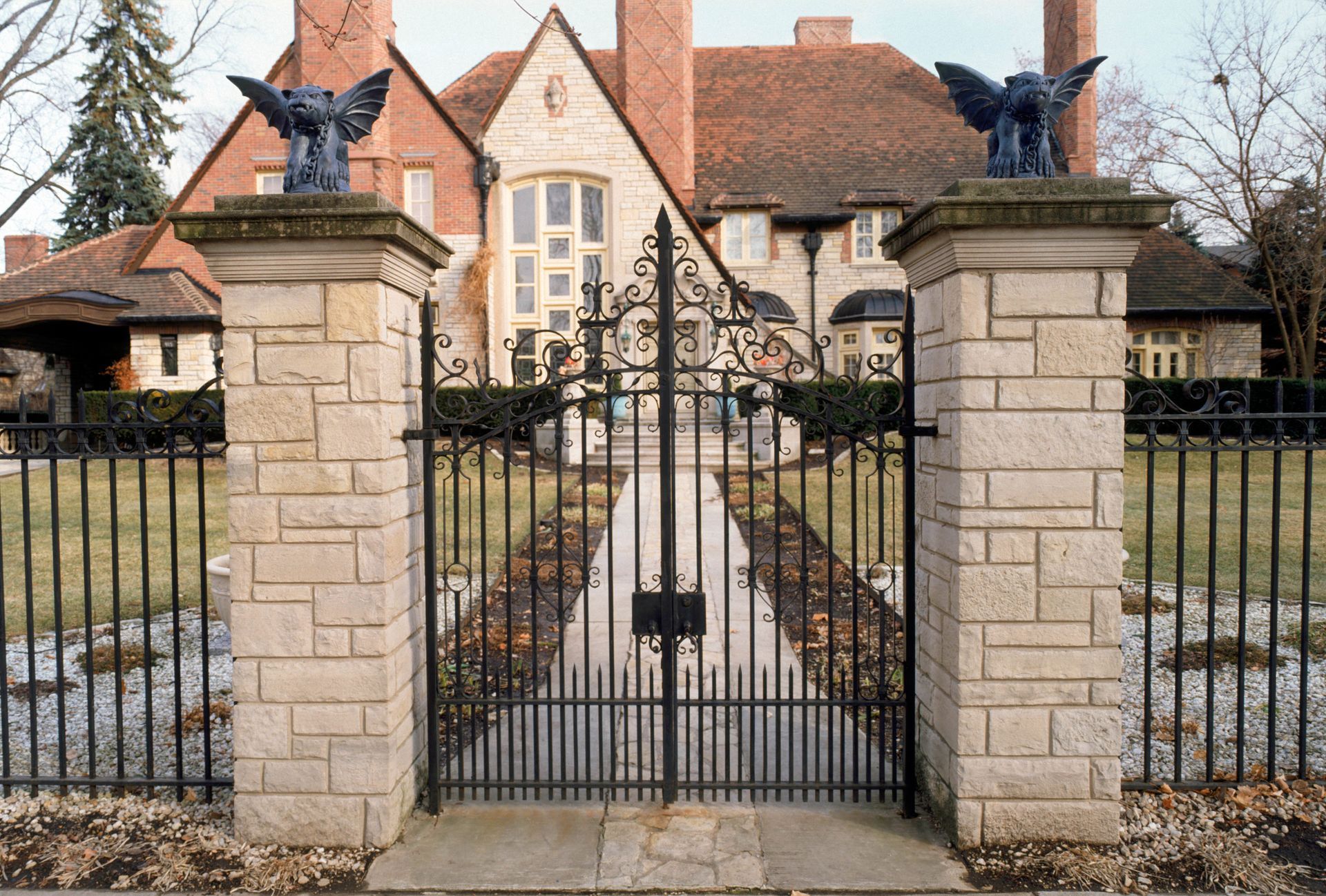 A stone wall with two pillars topped with gargoyle statues, enclosing a wrought-iron gate in front of a brick manor home.