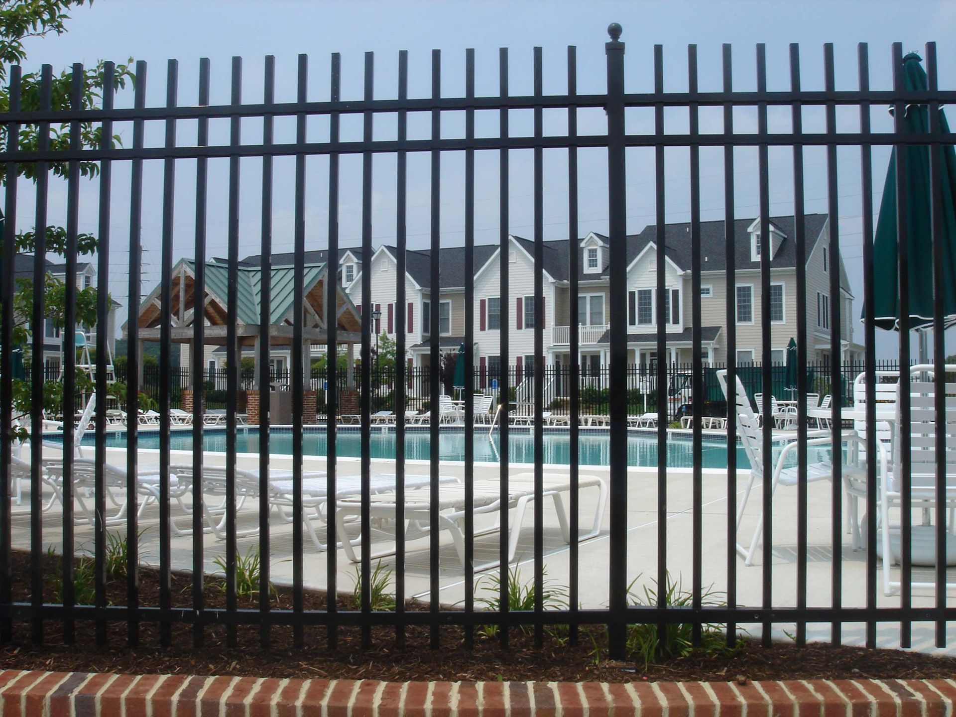 A black metal fence stands in the foreground, looking toward a swimming pool, lounge chairs, and a residential building.