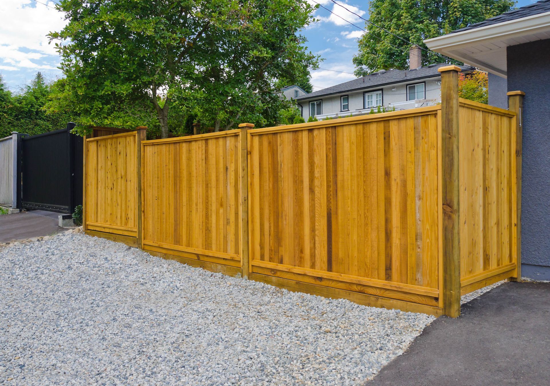 A modern wooden privacy fence installed between a gravel area and a paved driveway, with trees and a house in the background.