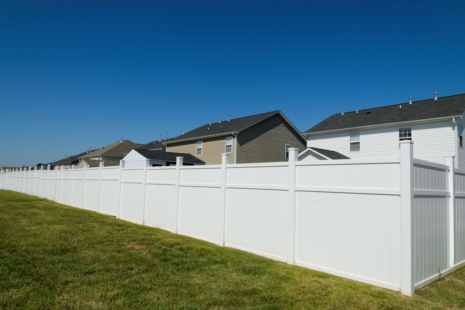 A long, white vinyl privacy fence runs along a green grass lawn in front of several suburban houses under a blue sky.