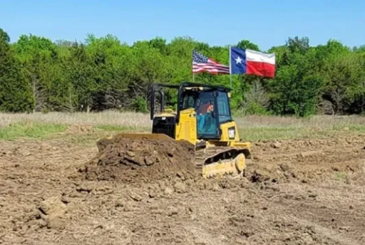A bulldozer is moving dirt in a field with a texas flag in the background.
