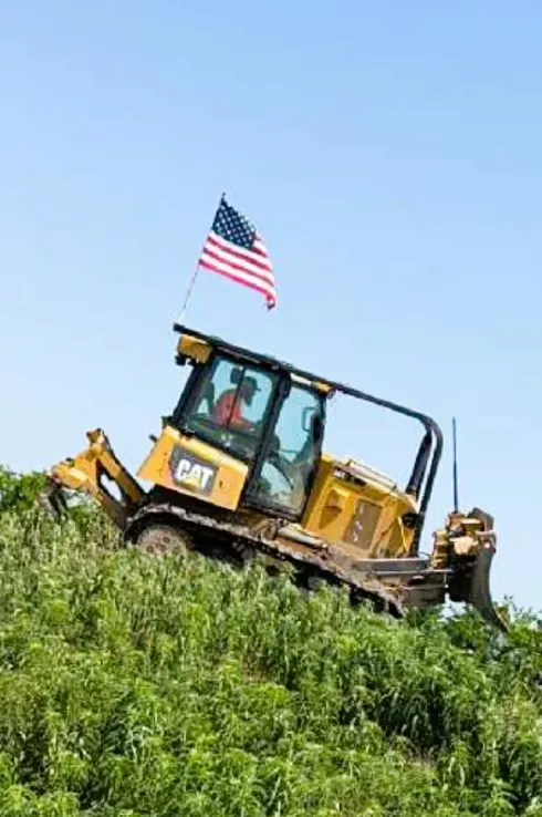 A bulldozer is driving down a hill with an american flag on top of it.