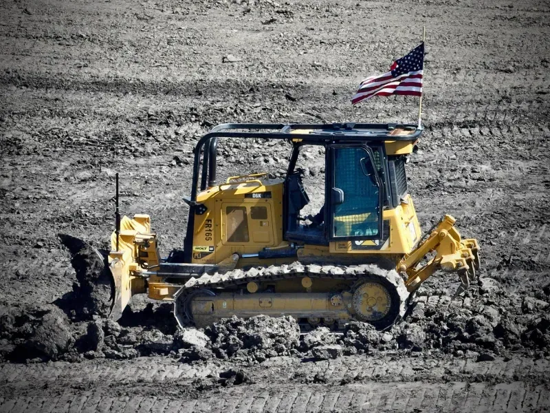 A bulldozer is driving through a muddy field with an american flag on top of it.