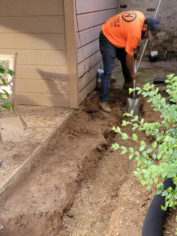 A man in an orange shirt is digging in the dirt with a shovel.