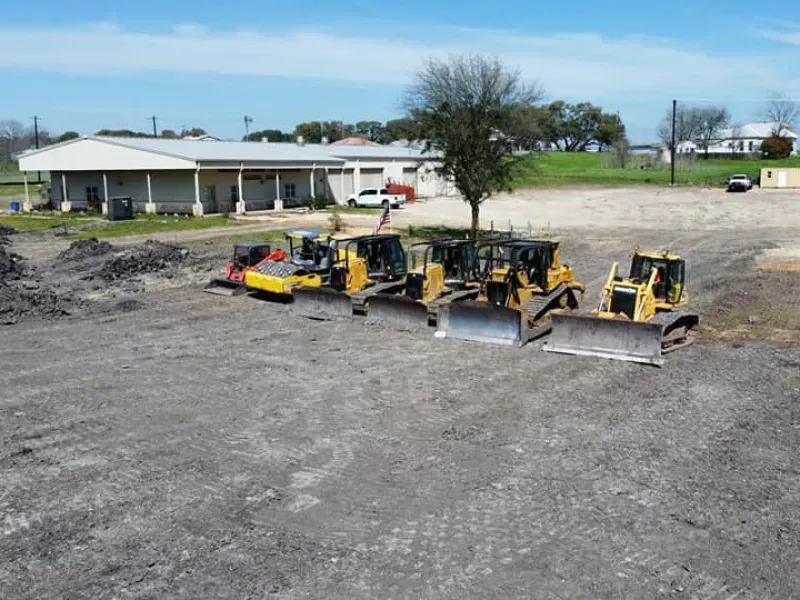A row of bulldozers are parked in a dirt field in front of a building.
