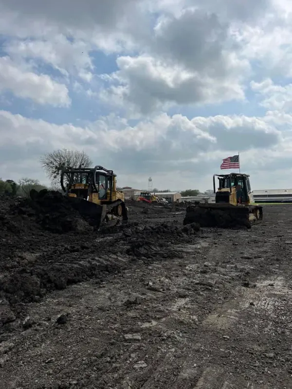 Two bulldozers are working in a dirt field with an american flag in the background.