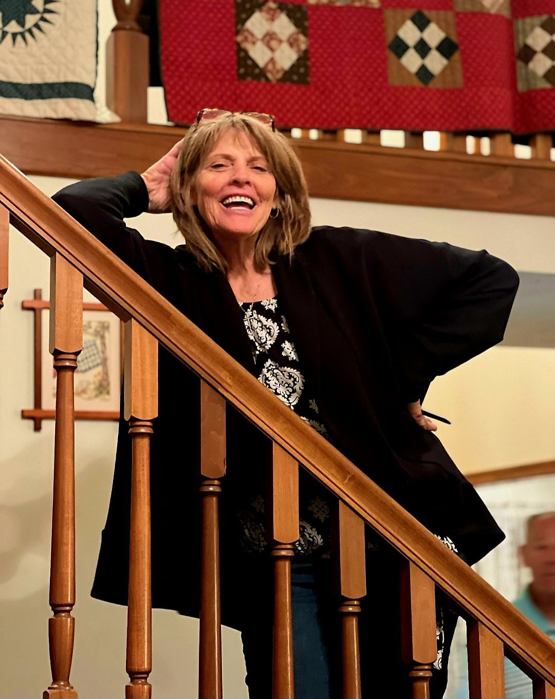 Woman smiling with hands behind head on a staircase, wearing a black cardigan and floral top.