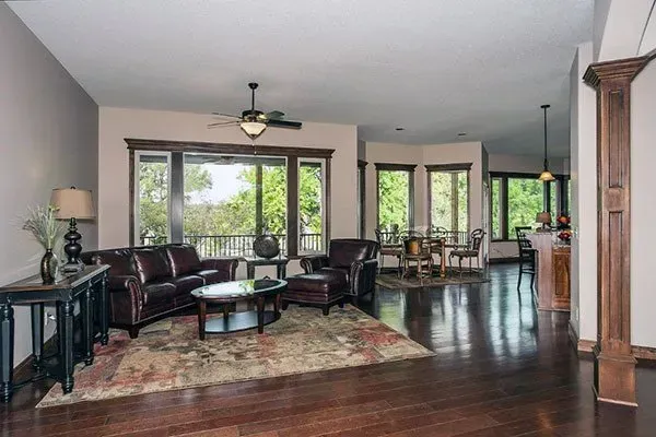 Spacious living room with dark wood floors, leather seating, a rug, and large windows overlooking greenery