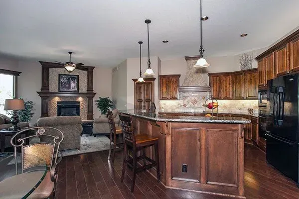 Warm kitchen with dark wood cabinets, granite island, pendant lights, and adjacent living room with fireplace.