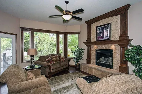 Bright living room with fireplace, bay windows, tan sofas, and a ceiling fan