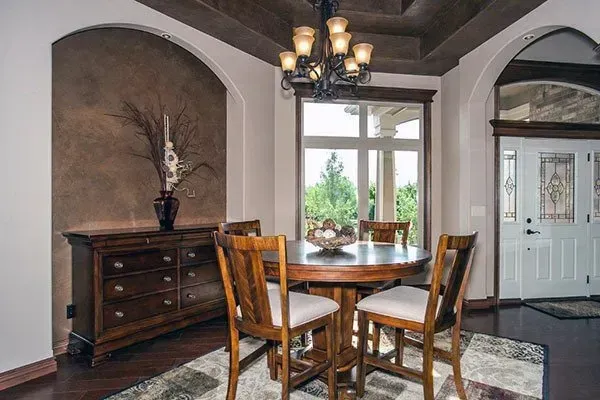 Dining nook with wooden table and chairs, dark dresser, arched ceiling, and chandelier by a bright window.