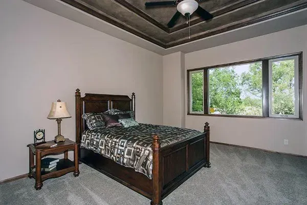 Bedroom with dark wood bed, nightstand lamp, gray carpet, and large windows