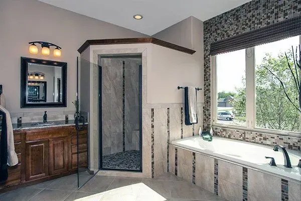 Luxury bathroom with glass shower, wood vanity, soaking tub, and large window overlooking greenery