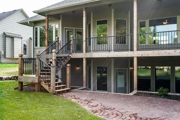 Backyard view of a raised house with a covered porch, stairs, and brick patio under the deck