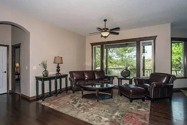 Living room with leather seating, patterned rug, ceiling fan, and large windows overlooking greenery