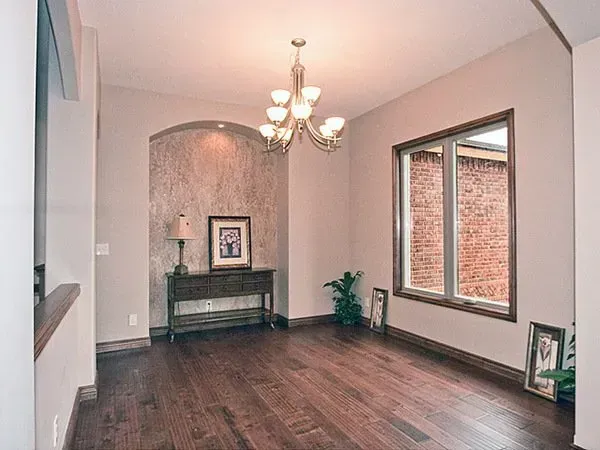 Empty living/dining room with dark wood floors, chandelier, and brick-accented window wall.