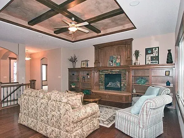 Living room with patterned sofa and armchair facing a wooden fireplace wall under a tray ceiling with fan