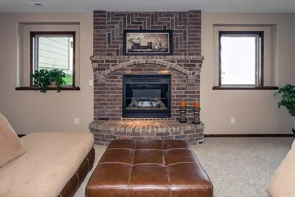 Living room with brick fireplace, two windows, beige sofa, and brown ottoman.