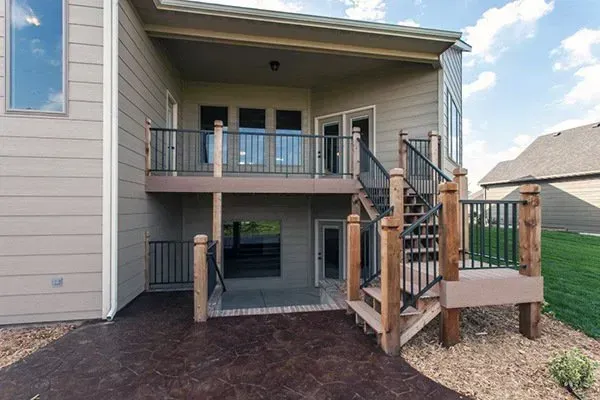 Backyard view of a raised patio with stairs, railing, and a covered balcony on a beige house