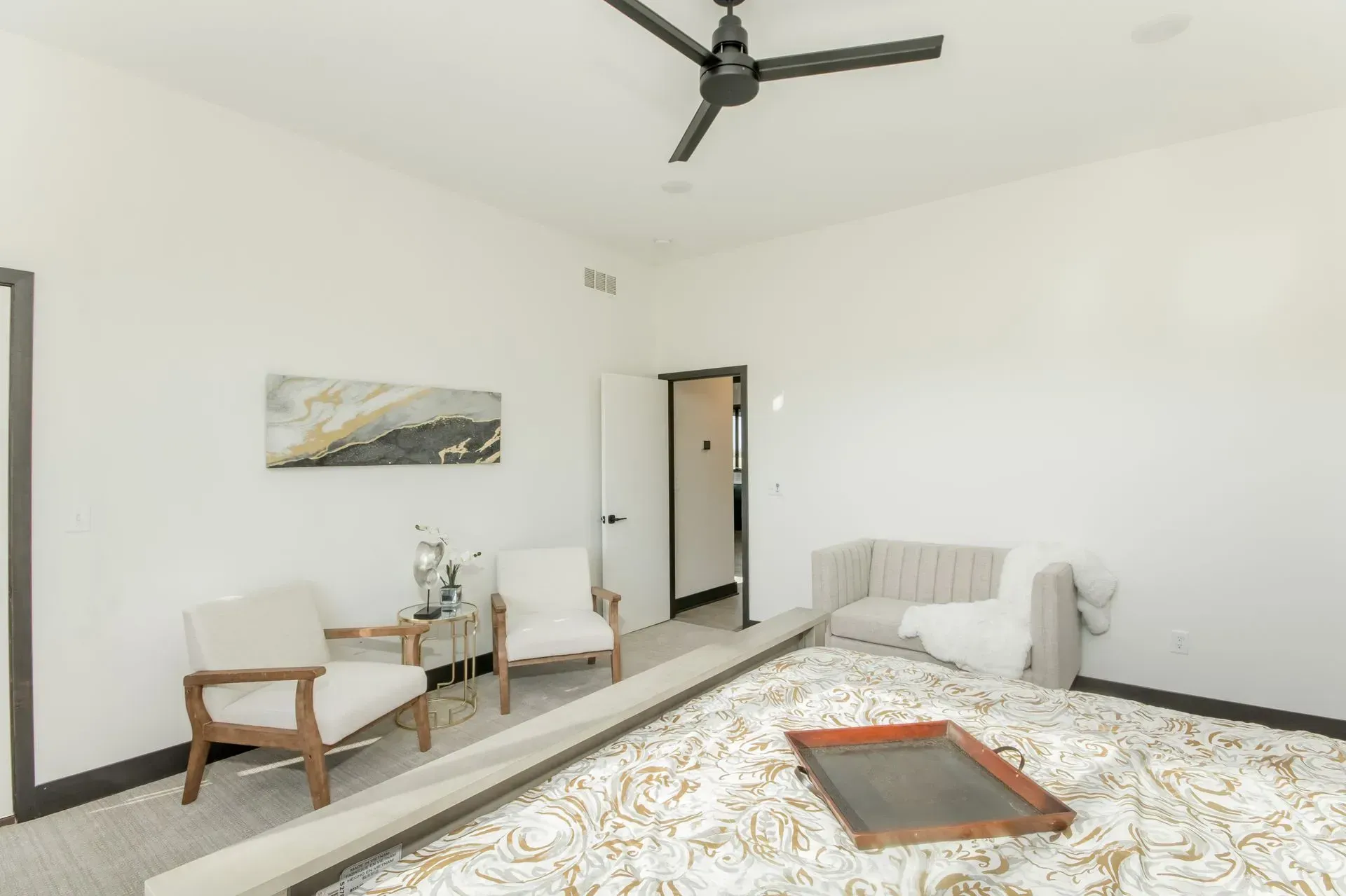 Bright bedroom with white walls, ceiling fan, bed, two chairs, and a small table near a doorway.