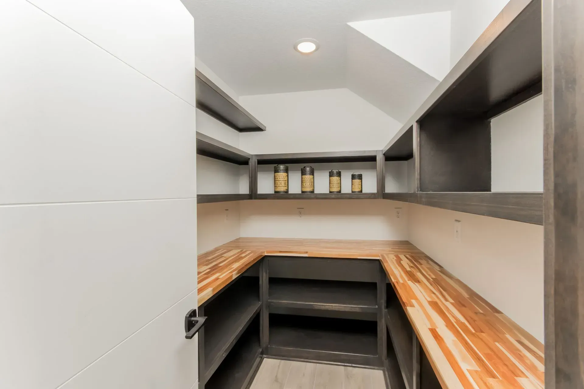 Small storage closet with L-shaped wooden shelves, gray walls, and bottles on the back shelf.