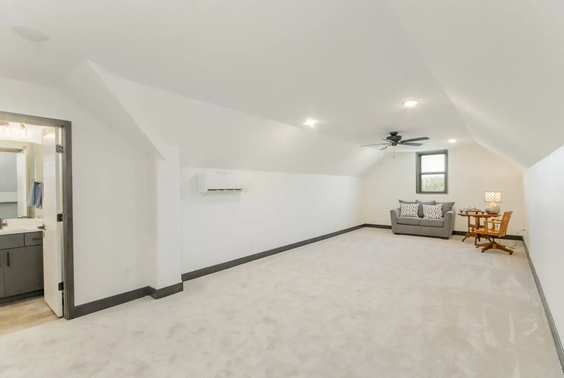 Bright finished attic room with white walls, carpet, a small seating area, and a wall-mounted air conditioner.