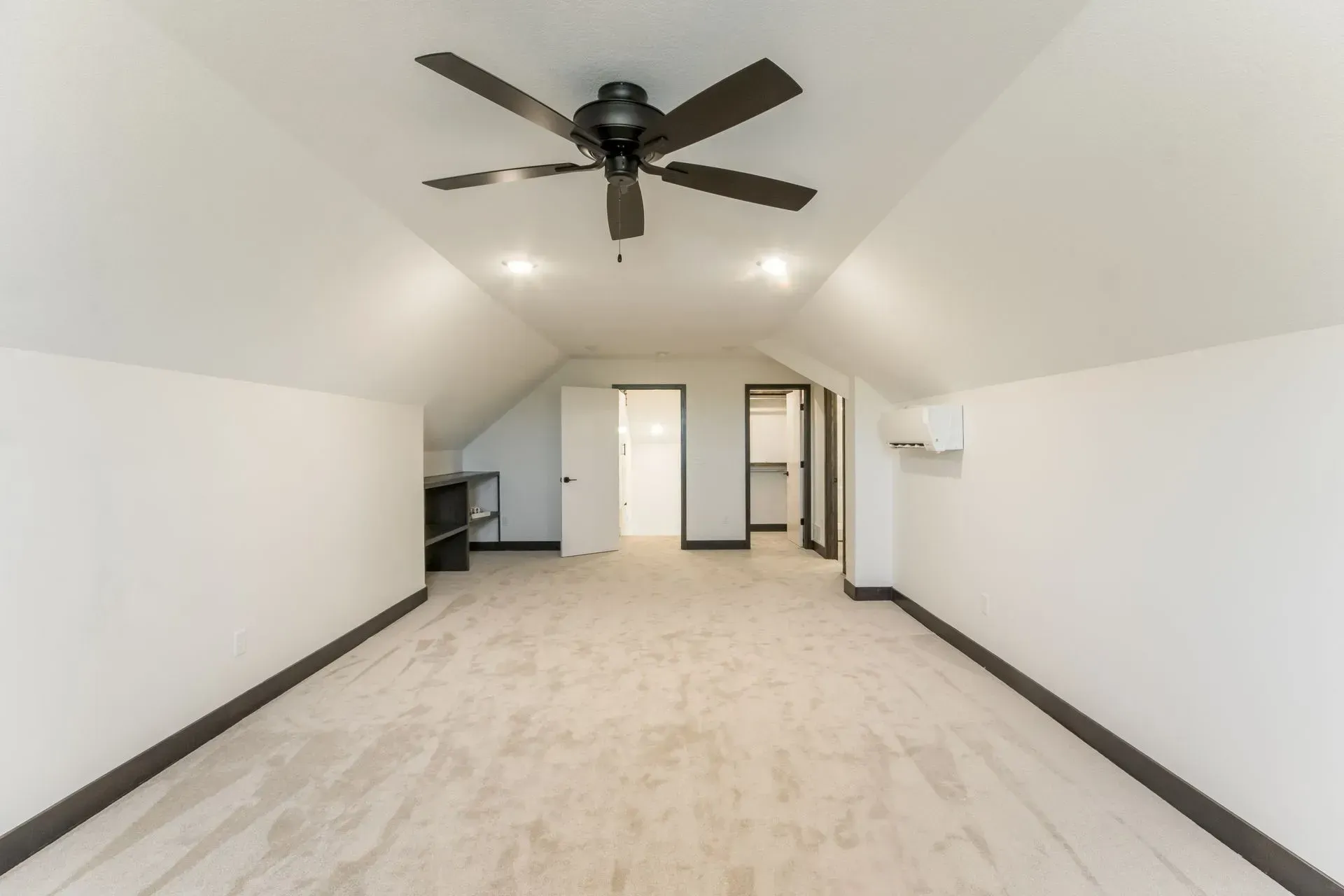 Spacious empty attic room with white walls, beige carpet, and a dark ceiling fan.