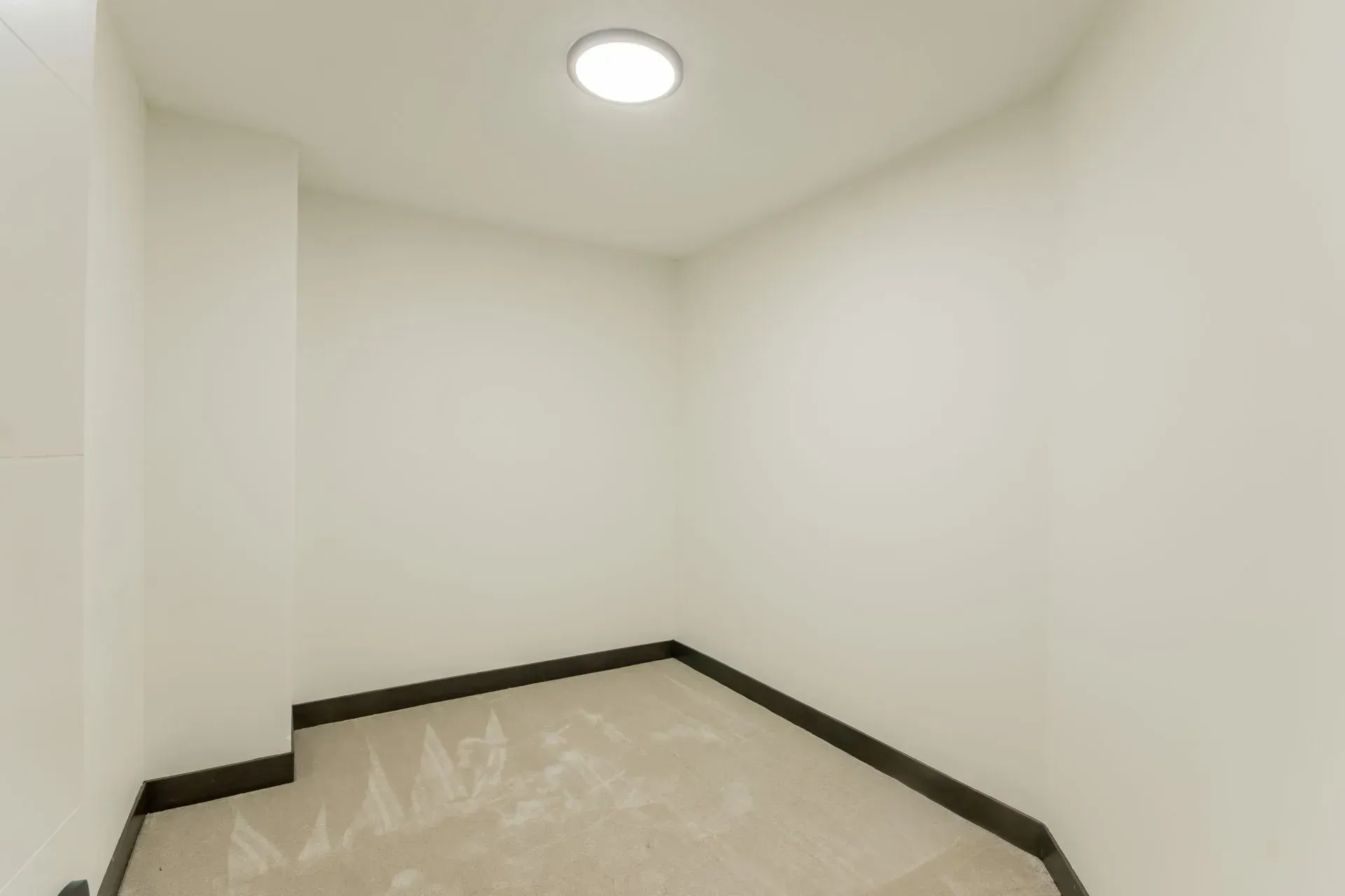 Empty white room with beige floor, dark baseboards, and a ceiling light.