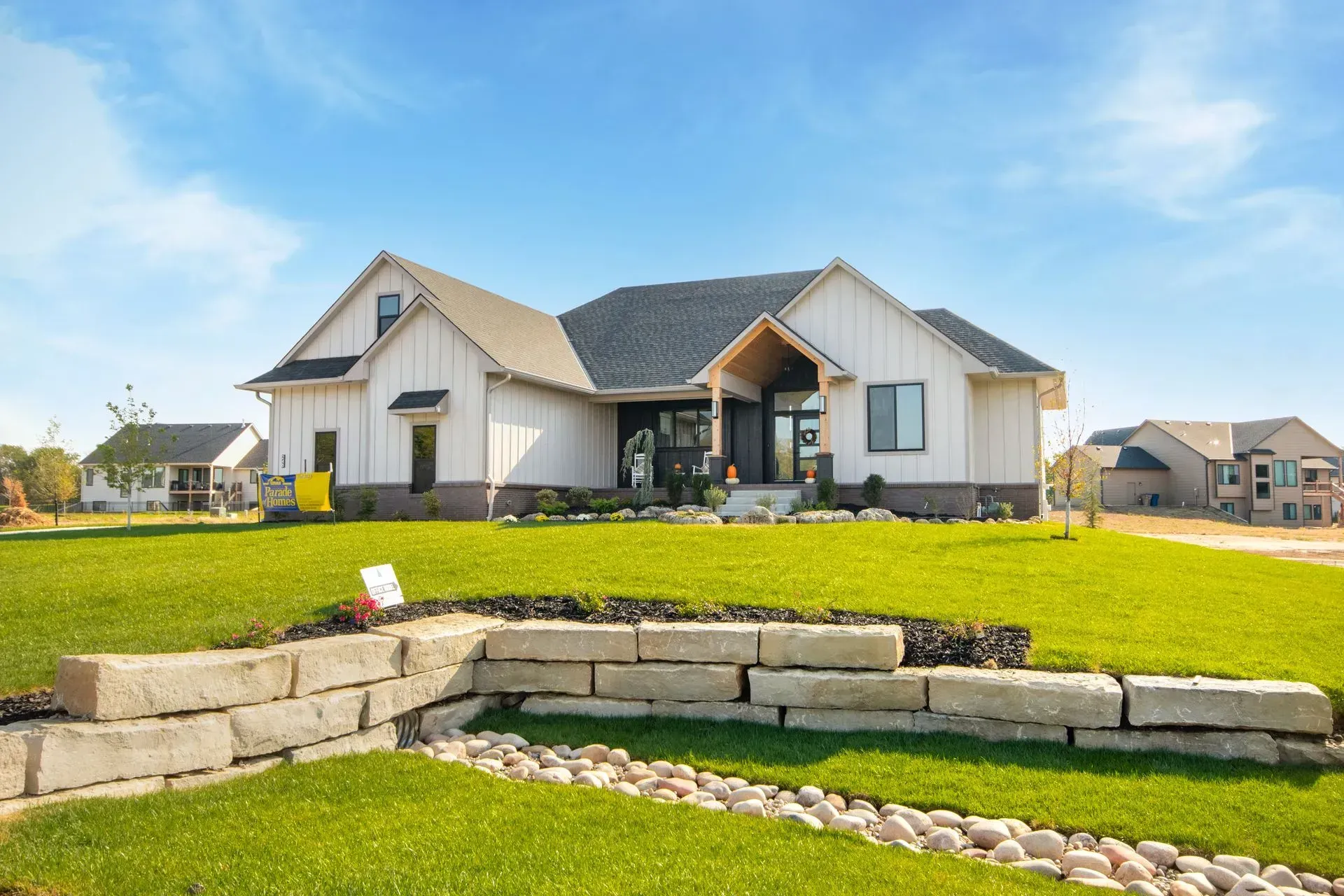 Single-story suburban house with a landscaped green lawn and stone retaining wall under a blue sky