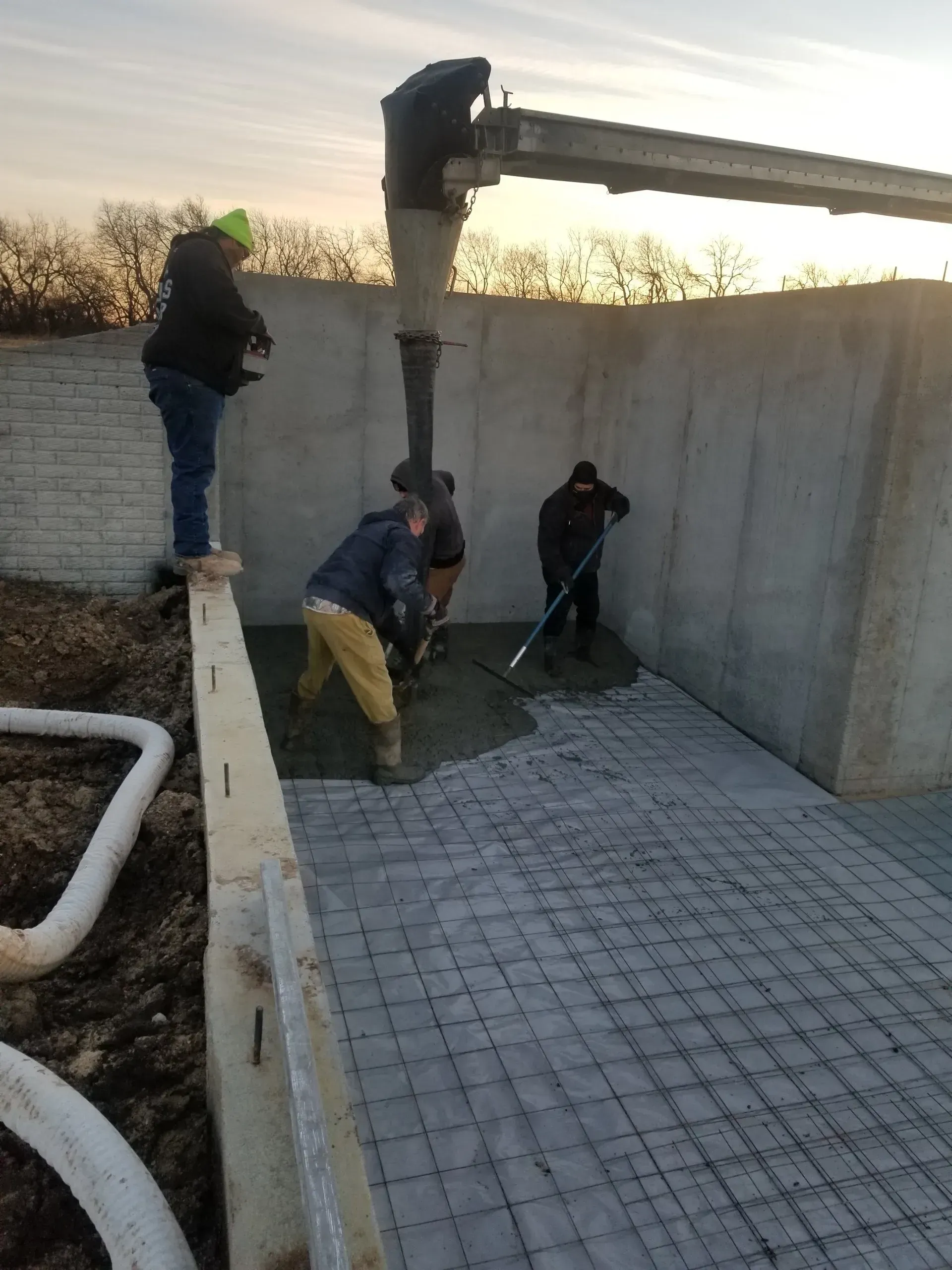 Workers pouring concrete into a large foundation wall form at sunset